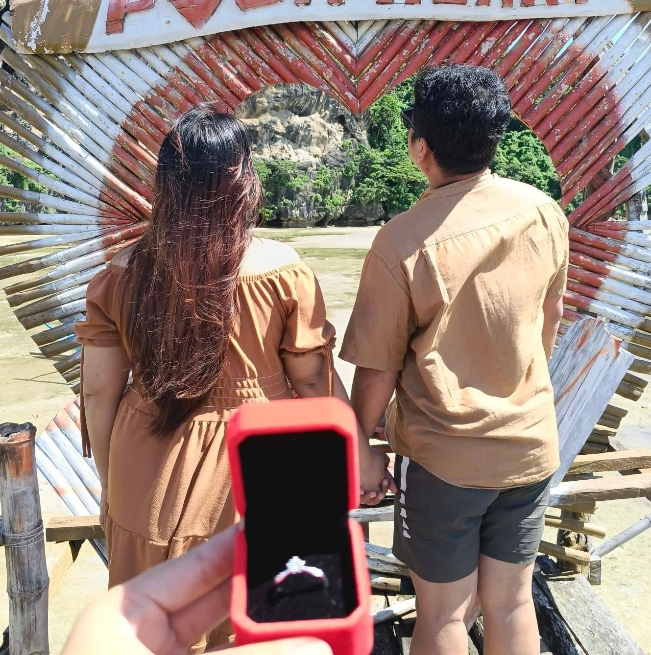 The couple holding hands in a heart-shaped frame, with a ring box in the foreground